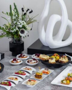 Assortiment de canapés gourmands sur une table de buffet avec bouquet de fleurs et sculpture blanche abstraite.